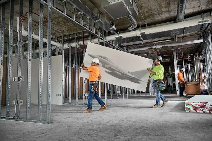 Workers carrying drywall through a commercial interior with steel-stud framing